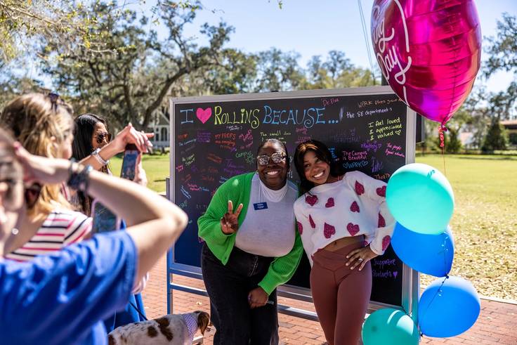Two women posing infront of a chalk board outside with blue and pink balloons and I Heart Rollins Because written on the board