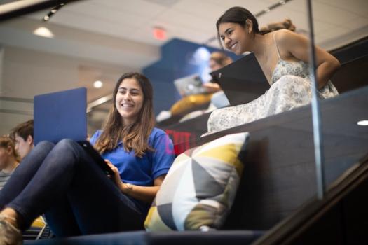 Two Rollins students chat in Kathleen W. Rollins Hall while holding their laptops.