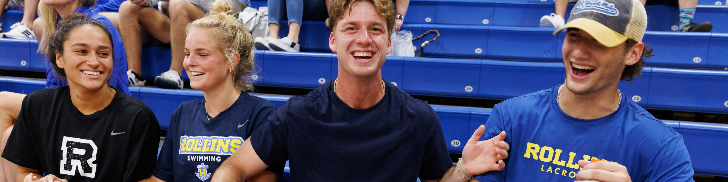 Four college students sitting on blue bleachers in Rollins athletics shirts