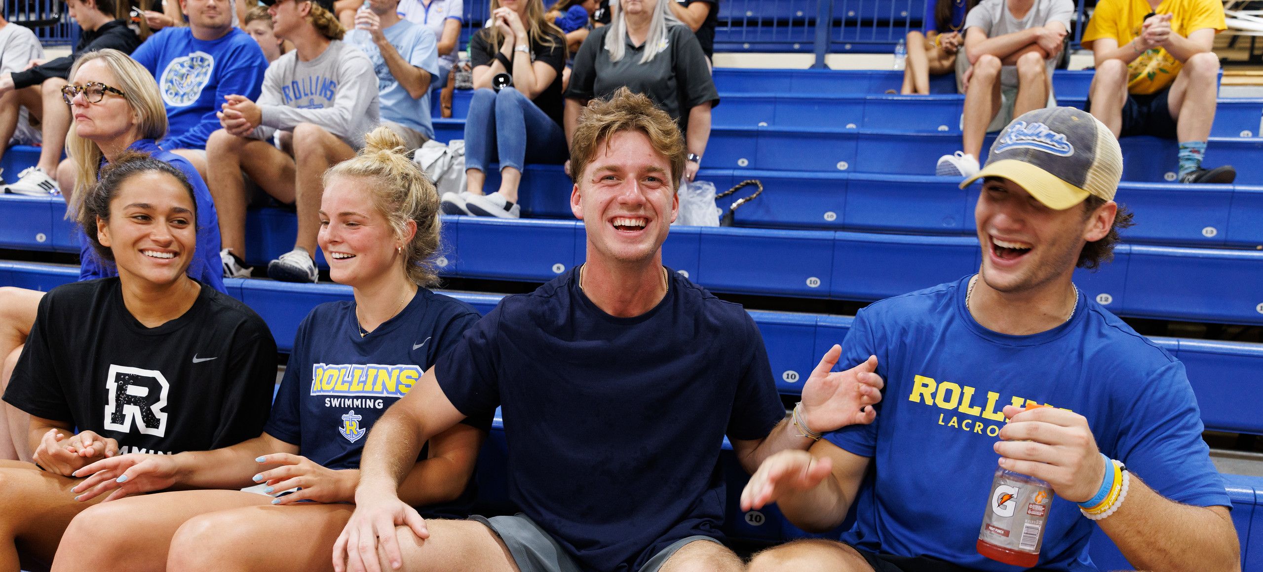 Four college students sitting on blue bleachers in Rollins athletics shirts