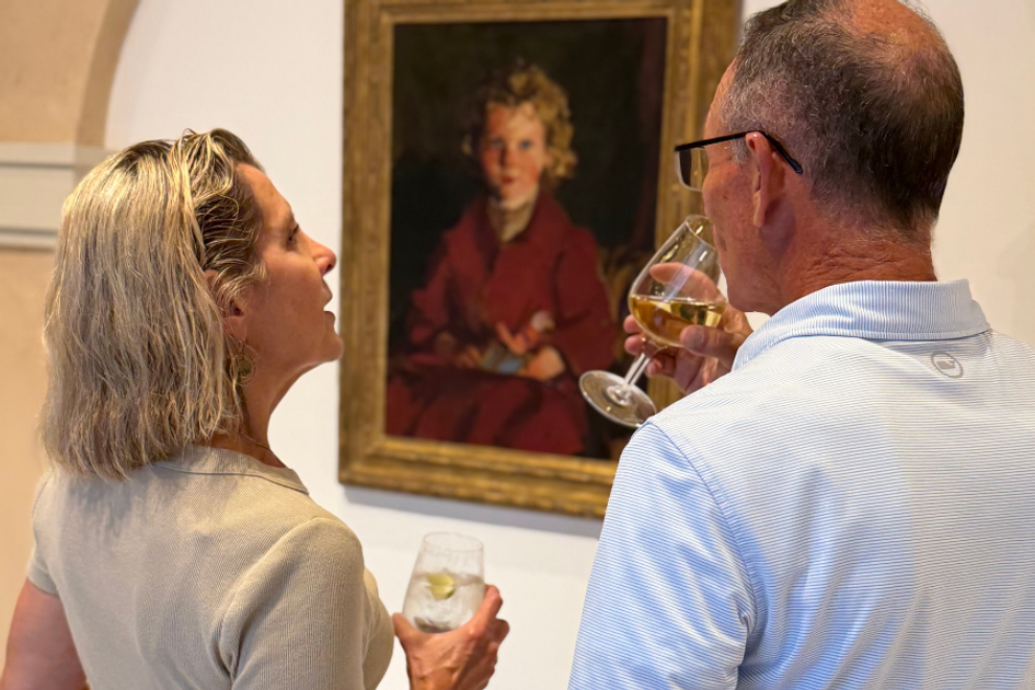Two people drinking wine while viewing a framed portrait in gallery.