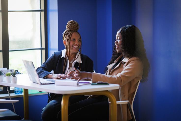 A Rollins alumna and a student sit together at a desk, smiling and talking while viewing a laptop.