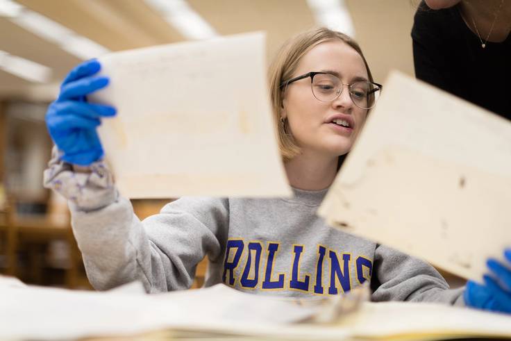A college student is holding up historical documents in each hand while wearing protective gloves.