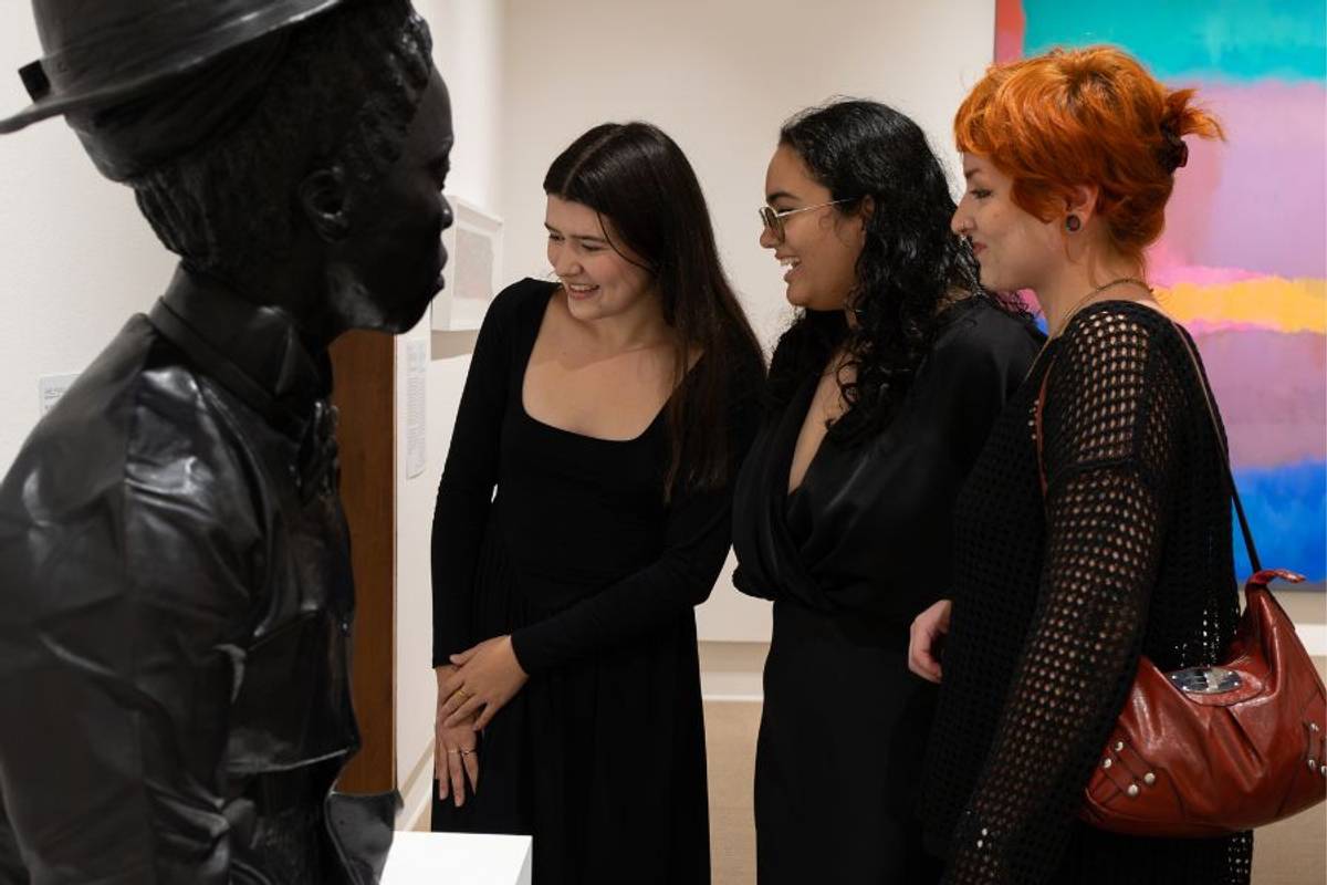 Three students stand in a gallery looking at a bust sculpture with a colorful painting displayed in the background