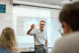 Political science professor speaking to class in front of a while board with notes and a Jeopardy projection.