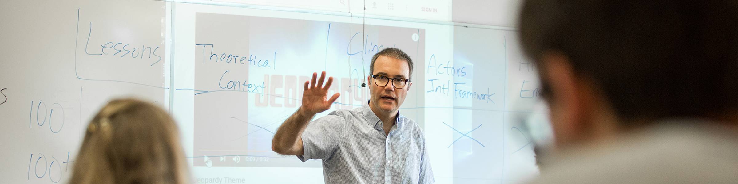 Political science professor speaking to class in front of a while board with notes and a Jeopardy projection.