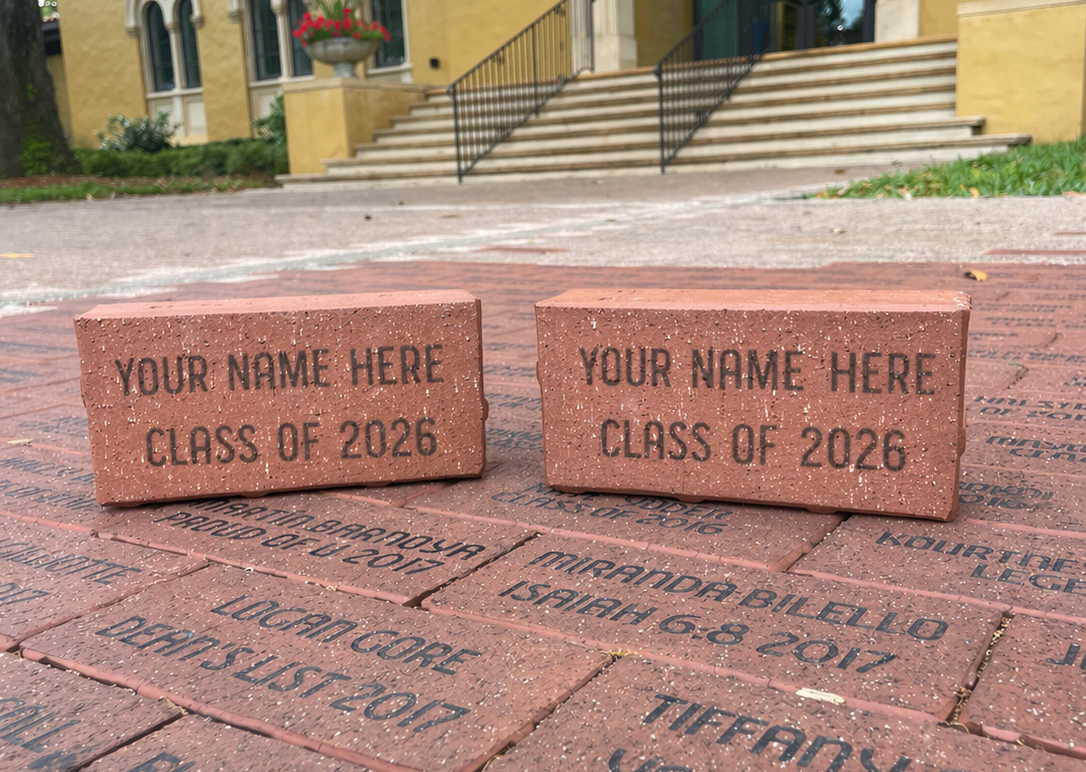 Two red bricks engraved with "YOUR NAME HERE CLASS OF 2026" rest on a pathway of personalized bricks in front of a building.