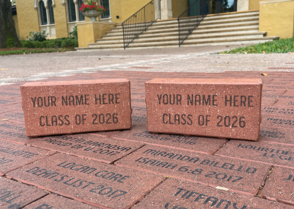 Two red bricks engraved with "YOUR NAME HERE CLASS OF 2026" rest on a pathway of personalized bricks in front of a building.