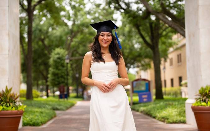 Sydney Boswell ‘25 in cap and gown smiling on a leafy campus walkway