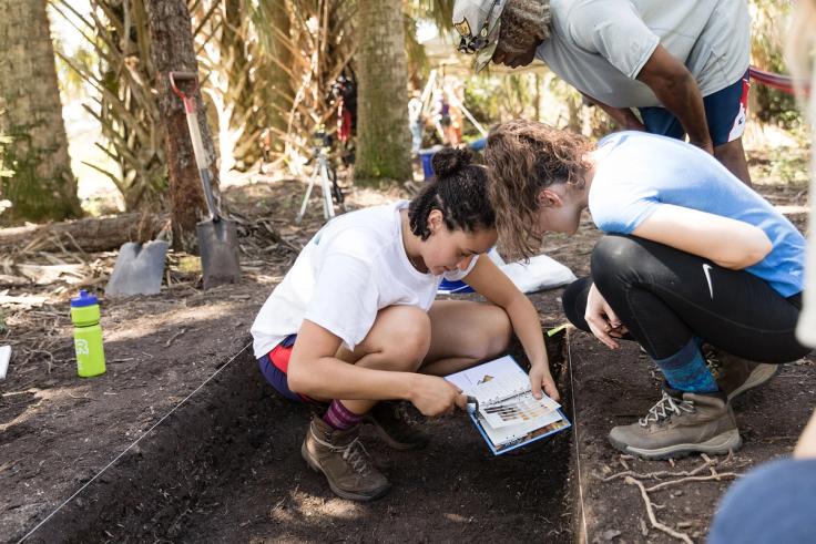 Anthropology students examine soil samples and notes at an outdoor dig site.