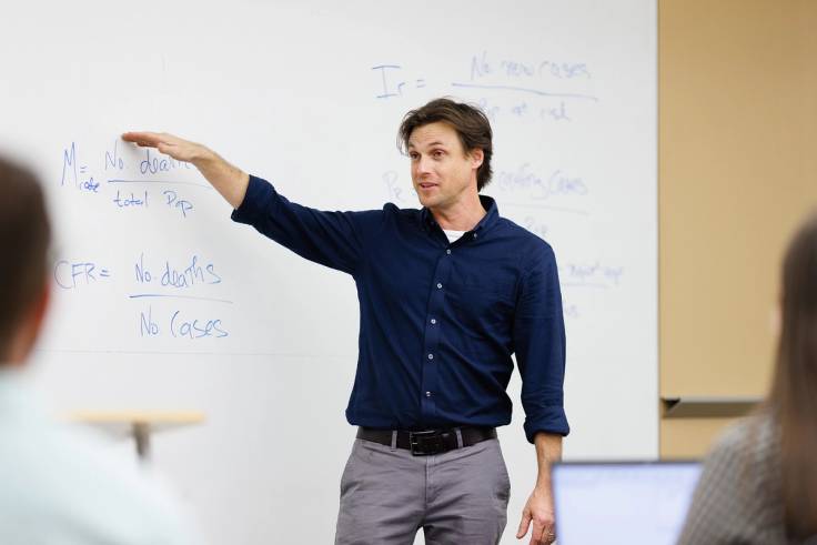 A public health professor explains a concept by pointing at a whiteboard.