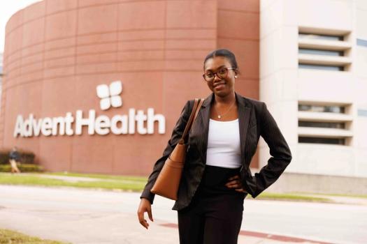 An intern poses in front of a hospital building with a large Advent Health logo on it.