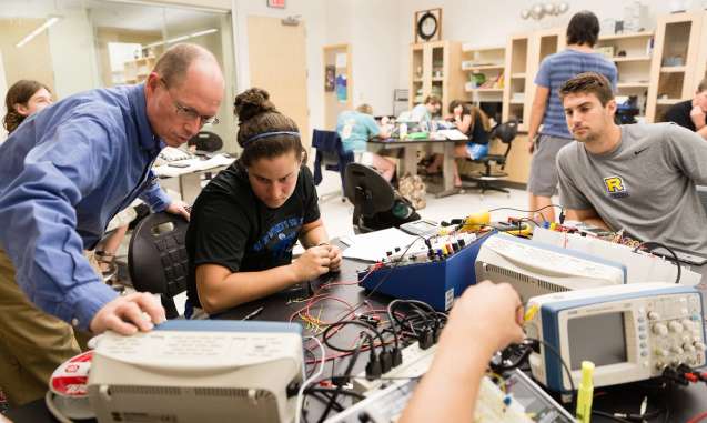 A physics professor is looking at a electrical reading with a couple of students in an electronics lab.