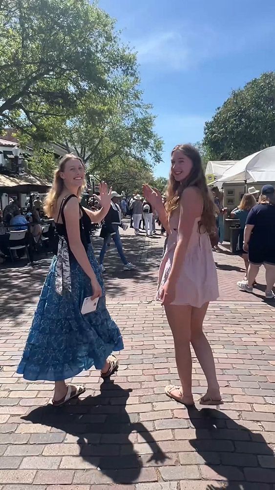 Two friends walking and waving in a sunny outdoor market street.