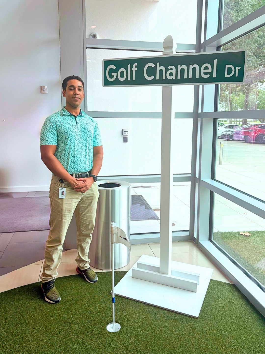 Student in a golf polo and khakis stands beside a "Golf Channel Dr" street sign on an indoor putting green.