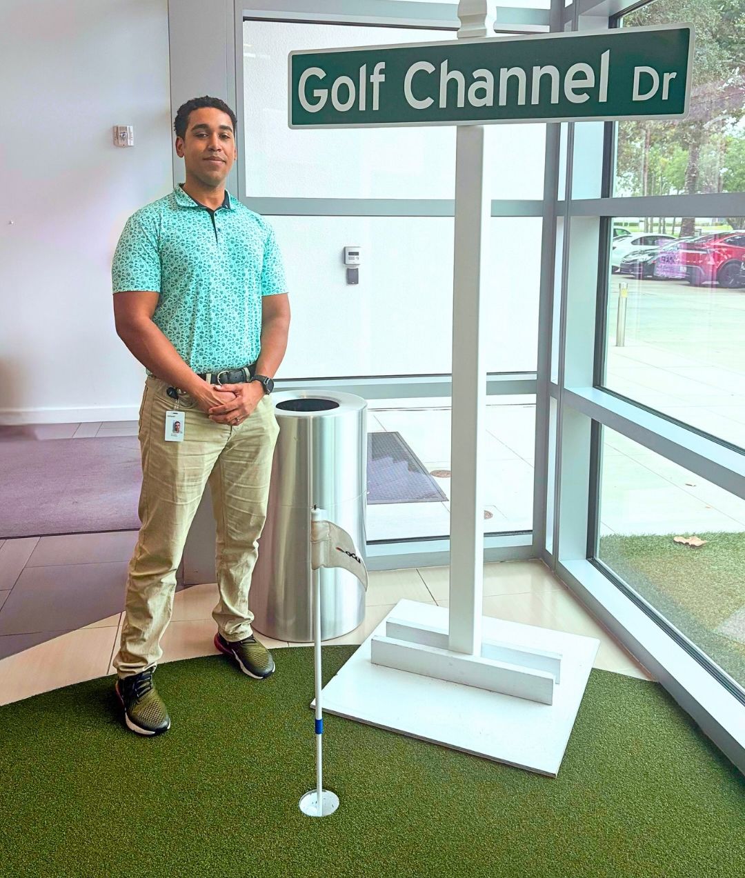 Student in a golf polo and khakis stands beside a "Golf Channel Dr" street sign on an indoor putting green.