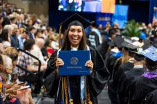 Student holds diploma dressed in cap and gown at Commencement ceremony with a large smile.