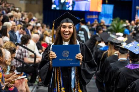 Student holds diploma dressed in cap and gown at Commencement ceremony with a large smile.
