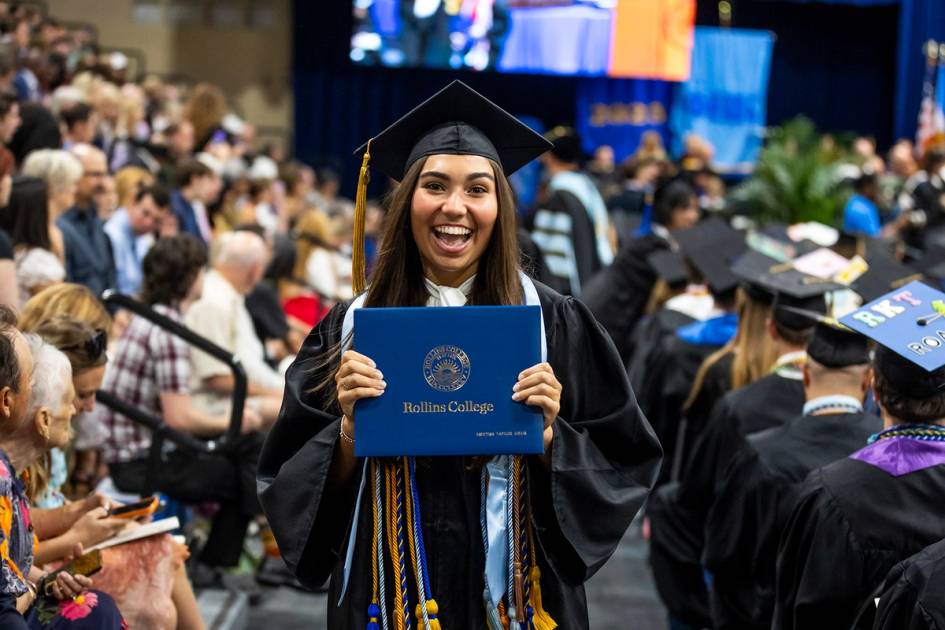 A beaming graduate in cap and gown holds up a "Rollins College" diploma case at her graduation.