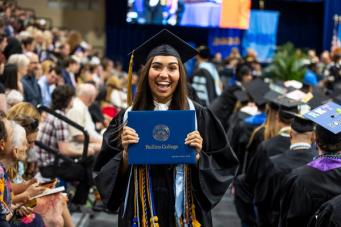 Student holds diploma dressed in cap and gown at Commencement ceremony with a large smile.