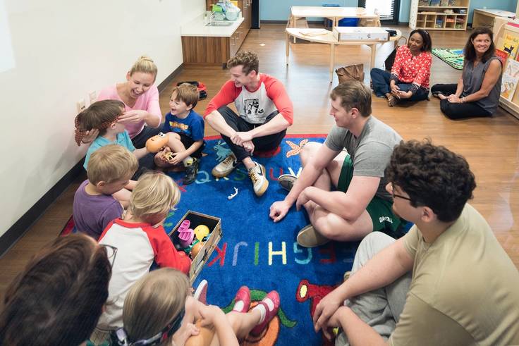 A preschool teacher and college students interact with small children in a classroom.