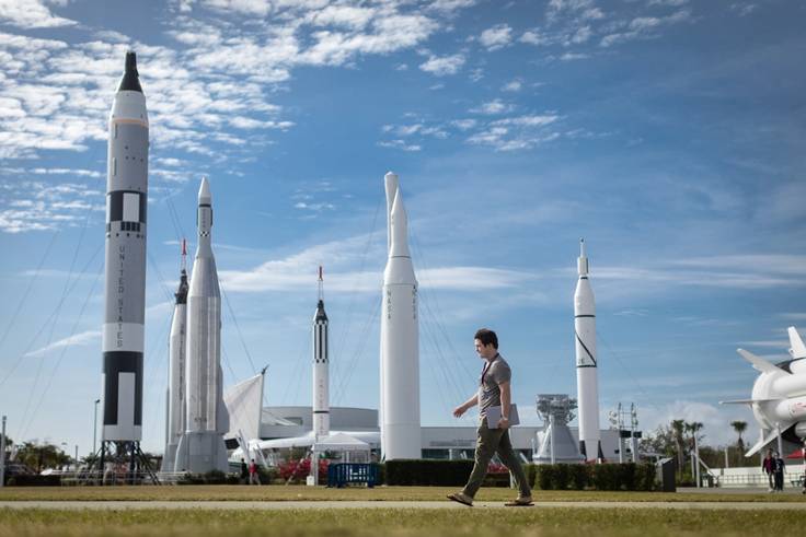 An intern at NASA's Kennedy Space Center carries his laptop with a half dozen rockets in the background.