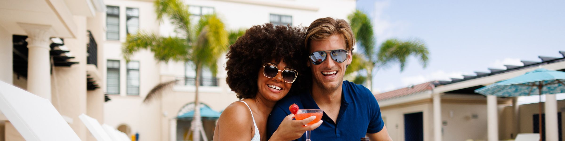 Hotel and pool in background, young couple sitting on poolside chair smiling and holding drinks.