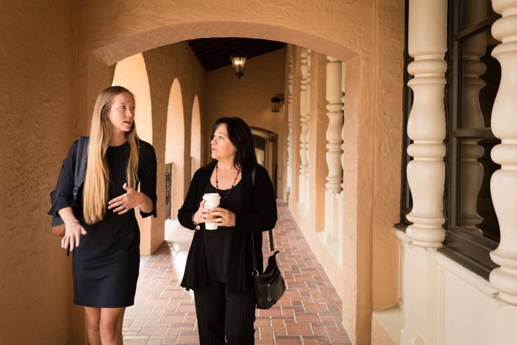 A student and her alumni mentor have a discussion while walking down a hallway on the Rollins College campus.
