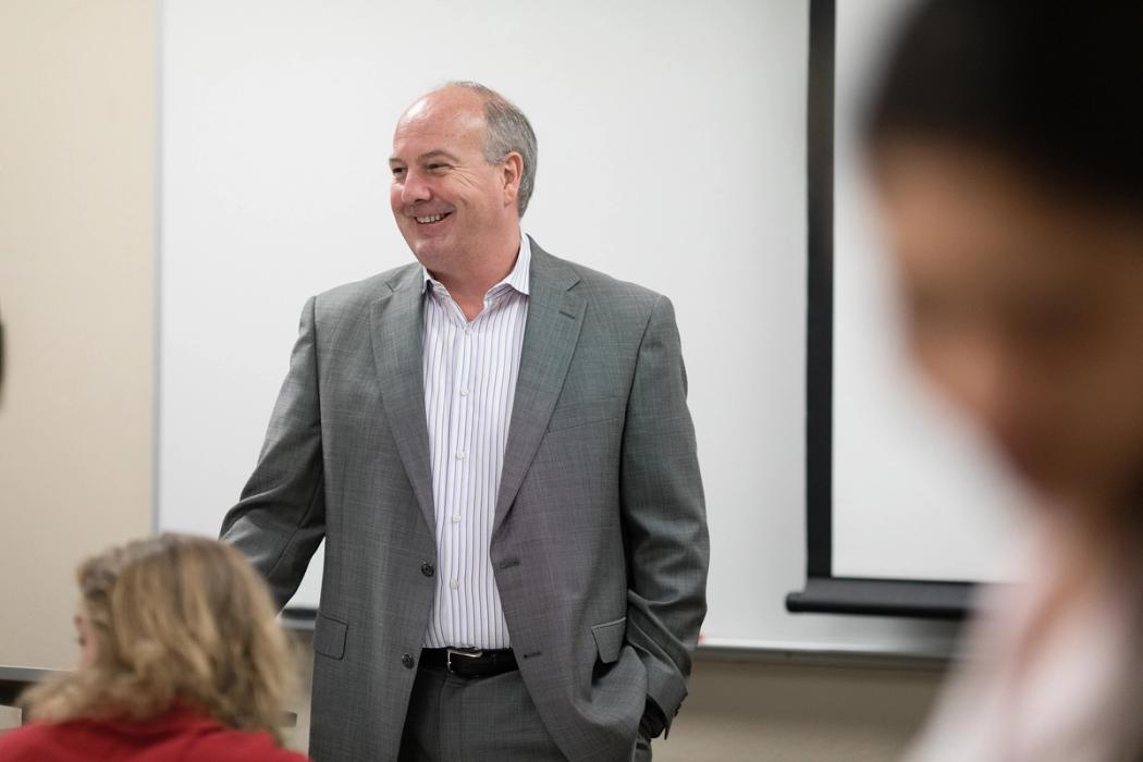 A business professor in a grey suit discusses a concept with a small group of students.