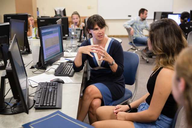 A college professor is sitting with her students in a computer lab explaining a problem.