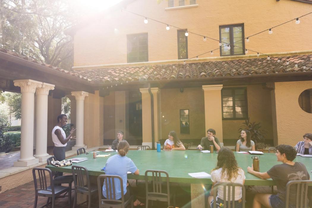 A professor leads a class discussion with students assembled around a large emerald table in an outdoor classroom on the Rollins College campus.