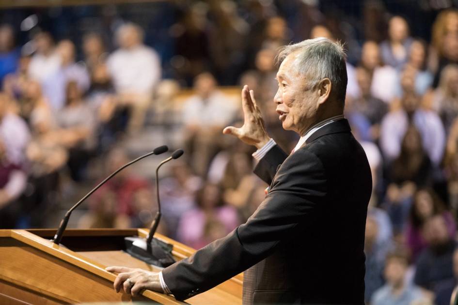 Speaker at podium addressing a large seated audience in auditorium.