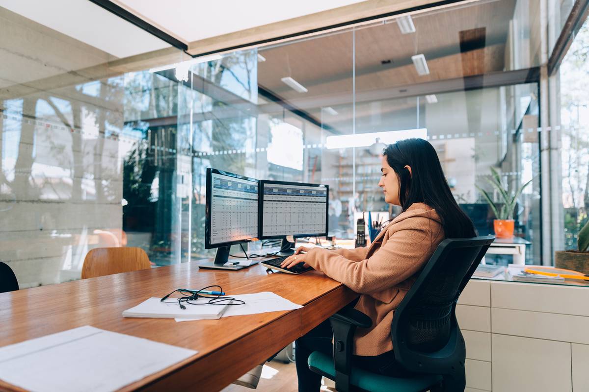 A financial analyst reviews data on a pair of computer screens in a glass-walled office.