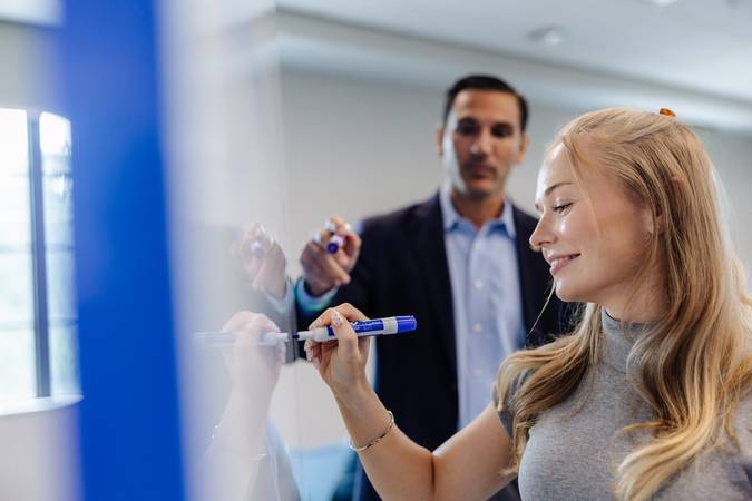 Smiling student writes on a whiteboard with a marker while a professor and classmates look on in a classroom.