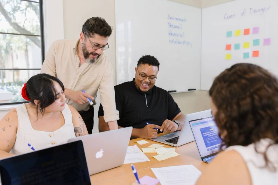 An instructor works closely with a small group of adult students gathered around laptops, collaborating on coursework in a classroom setting.