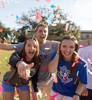 A group of Rollins students laugh while confetti falls; one student points at the camera.