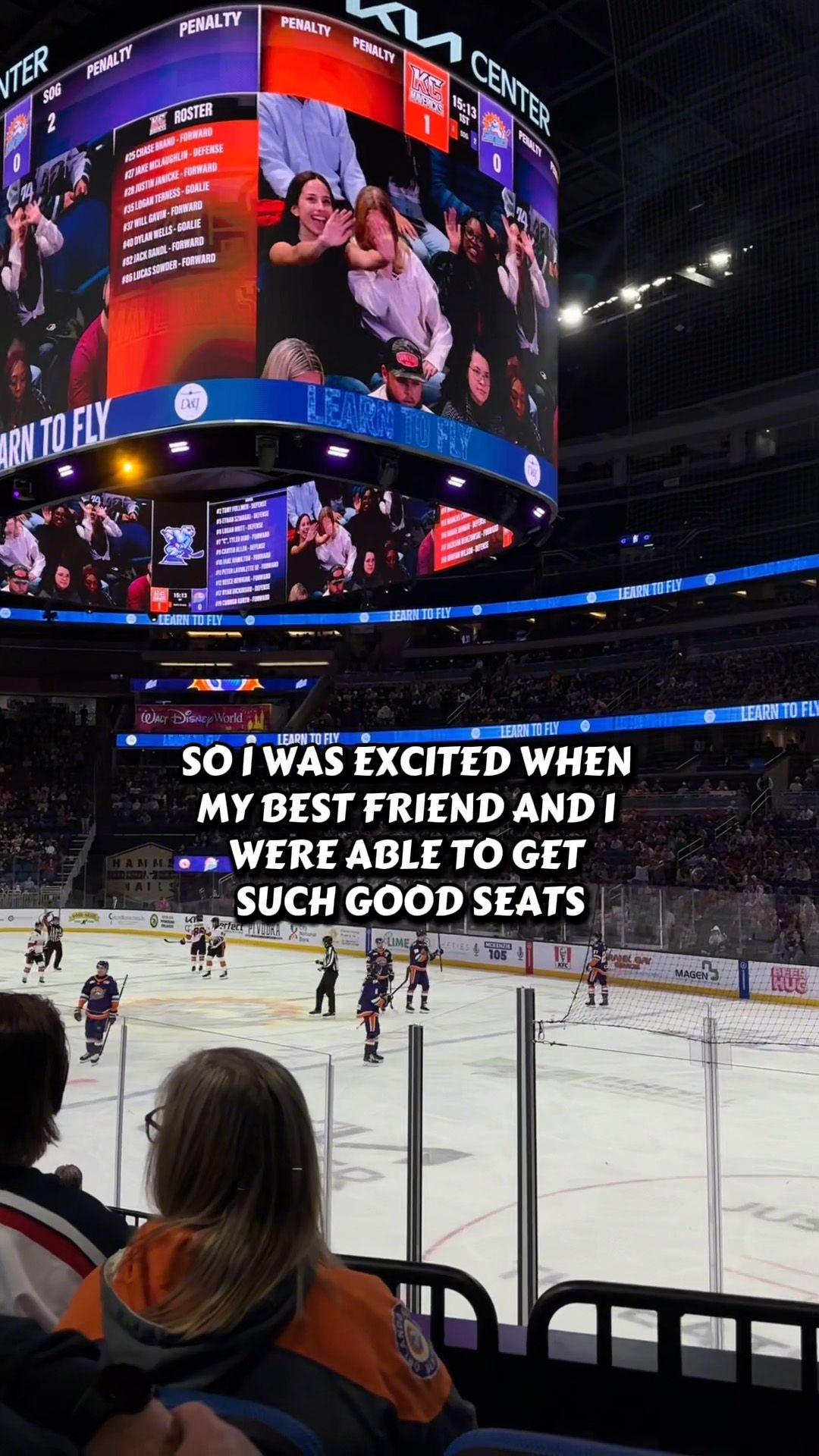 Fans enjoy close seats at an ice hockey game with jumbotron overhead.