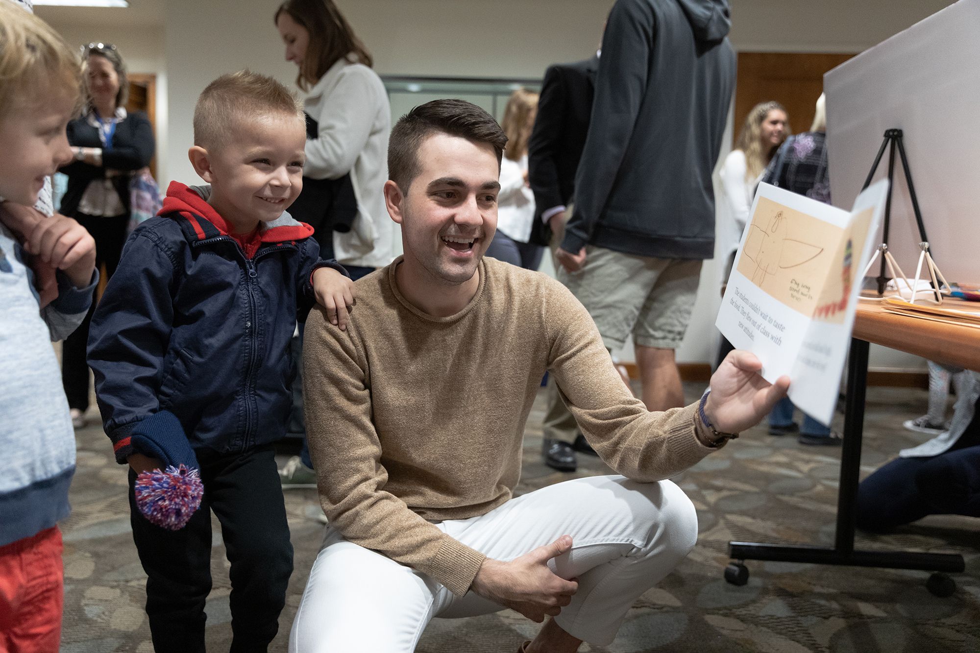 A student kneeling down reading a book to two children standing behind him.