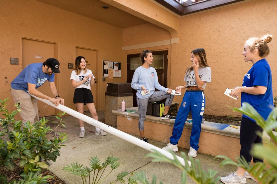 Professor Almond marking on a piece of pvc pipe as a group of girls are standing around holding staplers and talking.