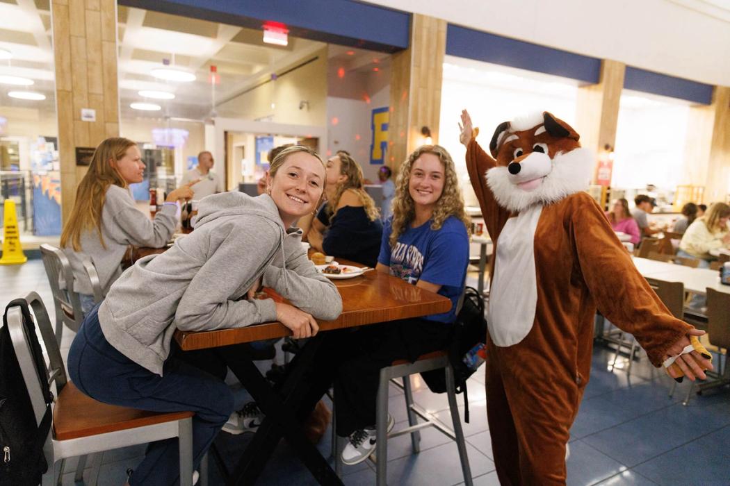 Two students pose with a fox mascot in the Rollins College dining hall.