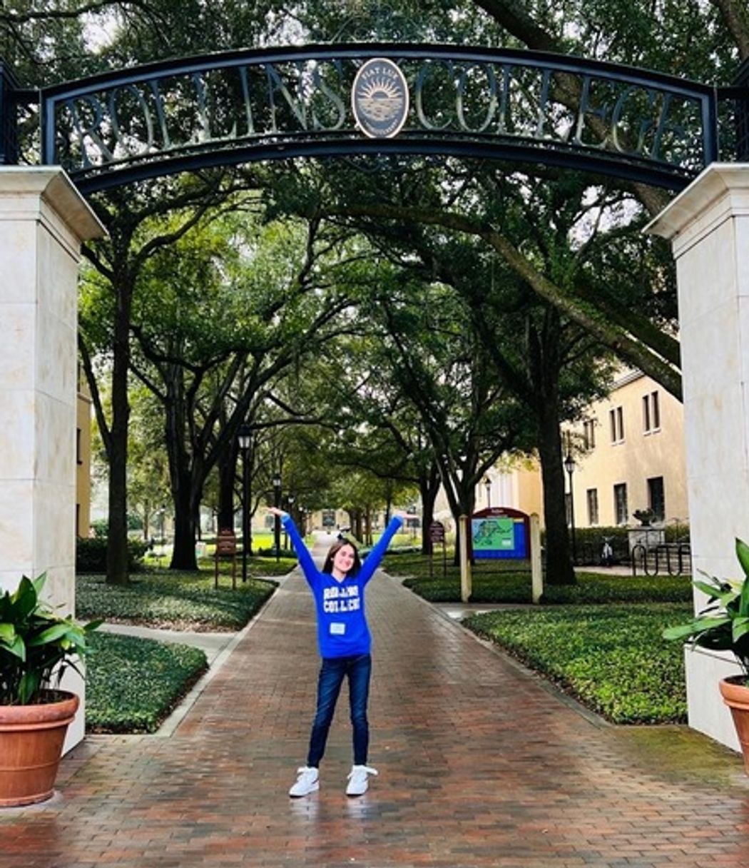 Student in a Rollins sweatshirt, excited to be attending Rollins College, standing in the archway entering campus