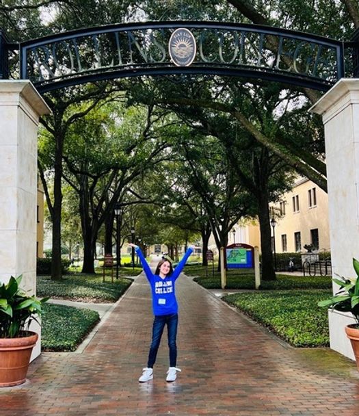 Student in a Rollins sweatshirt, excited to be attending Rollins College, standing in the archway entering campus