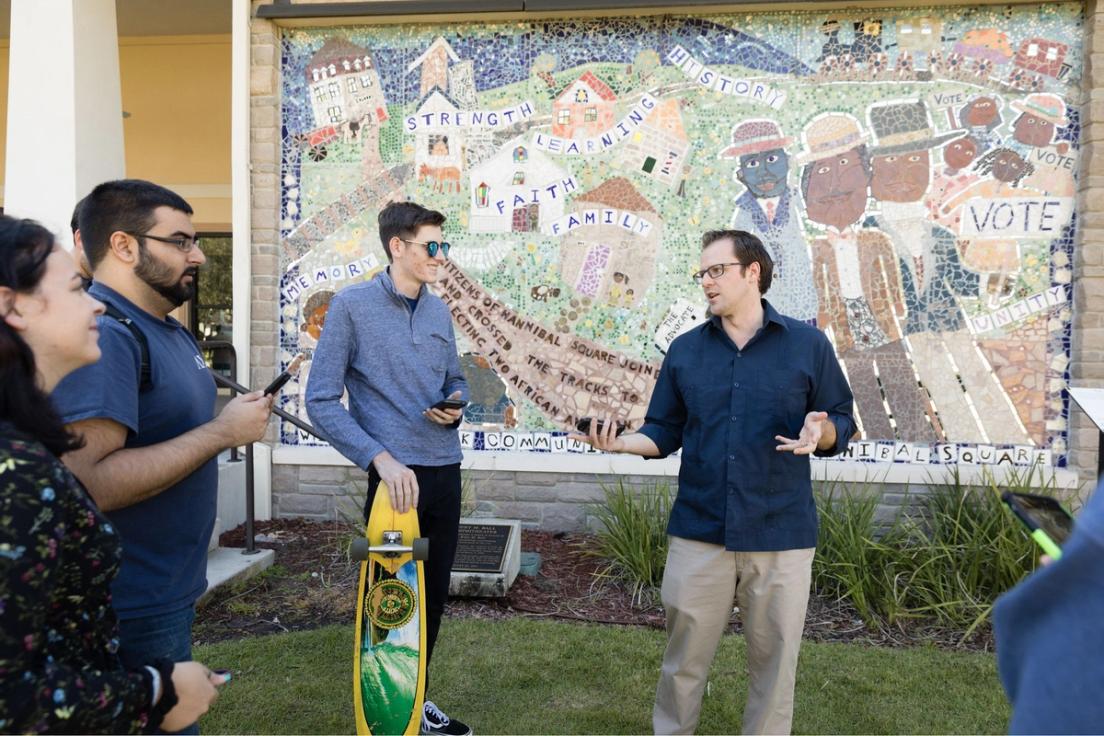 Professor Dan Myers standing in front of a wall mural at Hannibal Square talking to his students.