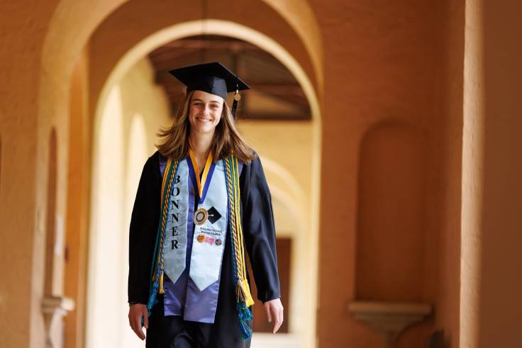 A college graduate walks on campus in cap and gown.