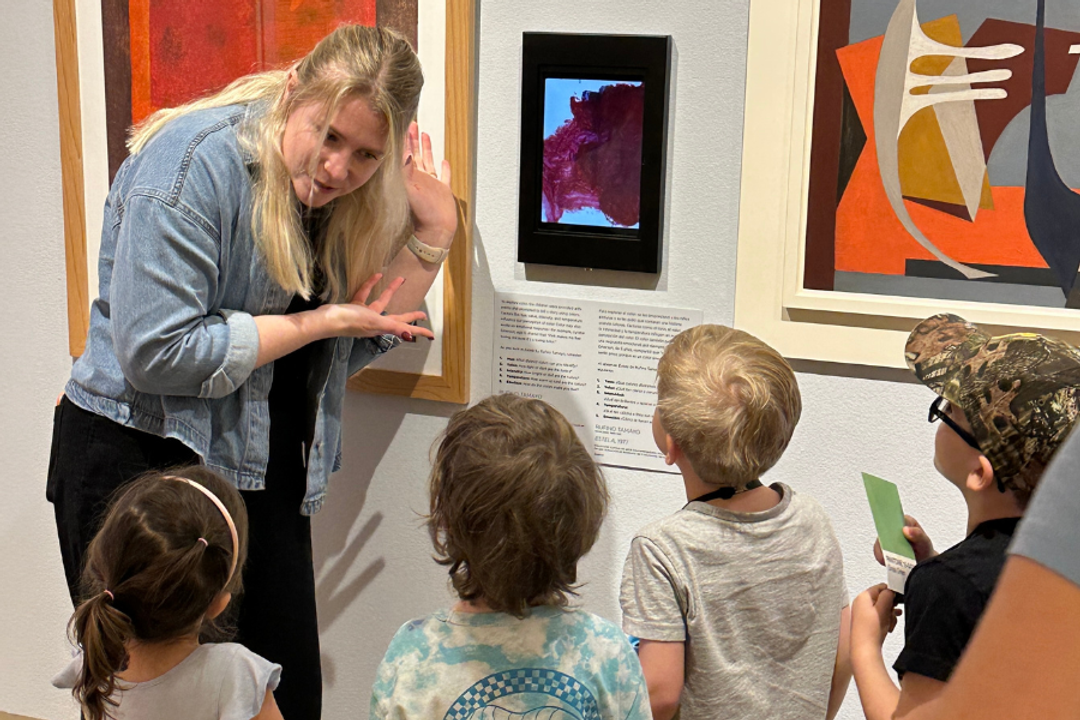 Children gather around an a woman who gestures to an artwork in a gallery