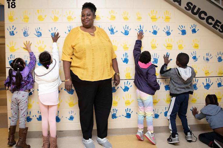 Elementary school paraprofessional stands alongside small children putting hand prints on a wall for decoration.