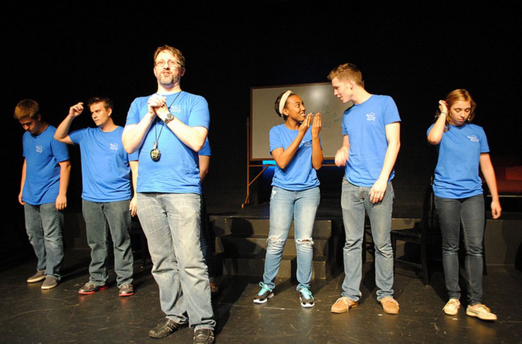 A group of students wearing matching blue t-shirts stand behind a faculty host with a stopwatch around his neck