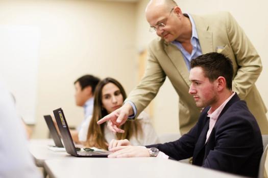 A business professor leans over a student to explain a concept on a computer screen.