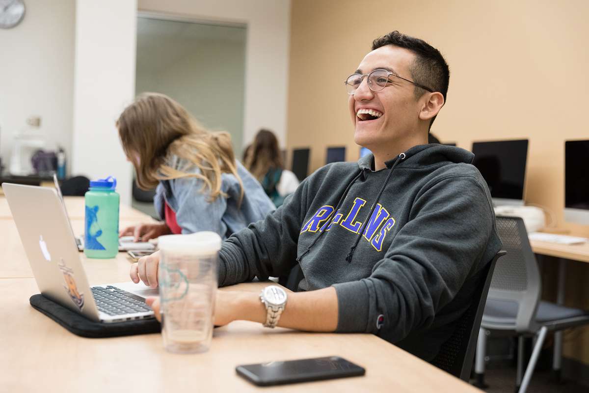 A male college student is sitting at a table with his laptop open laughing.
