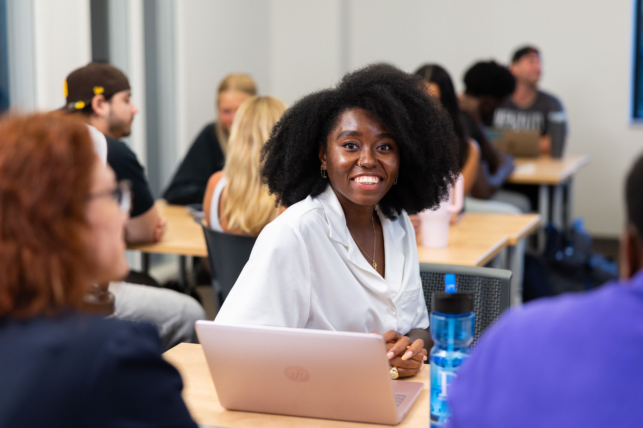 A smiling student in a white blouse sits at a desk with her laptop, talking with classmates during a small group discussion in a Rollins classroom.